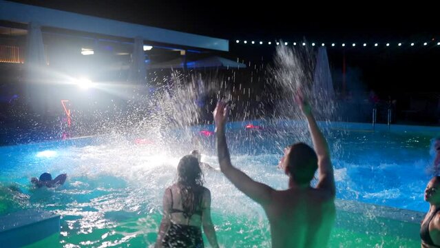 Friends have night pool party dancing in private villa swimming pool. Cheerful young people in swimwear jump and dive splashing water in luxury resort. Men and women hanging out in slow motion.