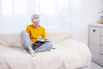 adult gray-haired woman is sitting at home on the couch and reading a book