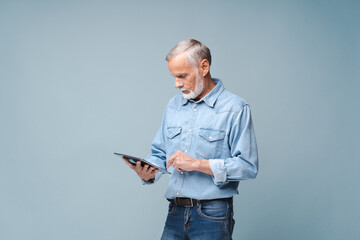 Photo of attractive senior man dressed in formal denim shirt using tablet computer isolated over blue background, touches pad screen, necessary tool for productivity, finds inspiration online