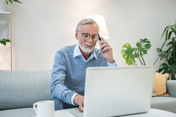 Cheerful senior man has phone conversation, chatting on the smartphone working with a laptop remotely from home. Waist up portrait view