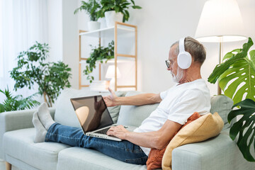 Grey haired mature man wearing headphones sitting at the couch and studying at his laptop at home. Retirement people concept