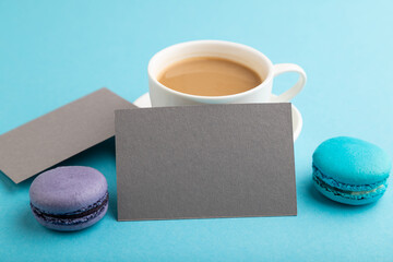 Obraz premium Gray paper business card mockup with blue and violet macaroons and cup of coffee on blue pastel background. side view, copy space.