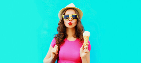 Portrait of happy young woman with ice cream blowing her lips wearing summer straw hat on blue...