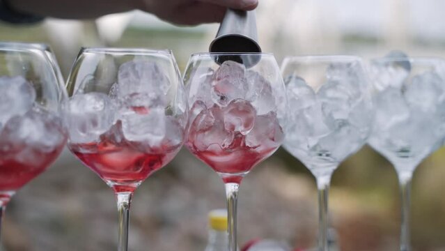 Bartender adds one part red drink from metal jigger to glasses with ice cubes standing on bar counter in open air. Preparation of refreshing drink in glasses standing in row.