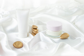 Hand cream tube and glass jar with empty space for label on white satin background. Cosmetic product photography backdrop