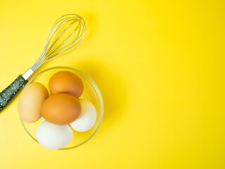 Top view of whisk and chicken eggs in glass bowl on yellow background,cooking concept