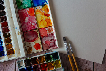 Palette of watercolor paints, blank paper and brushes on wooden background.  Artist's work space. Copy space. Selective focus.