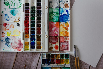 Naklejka premium Palette of watercolor paints, blank paper and brushes on wooden background. Artist's work space. Copy space. Selective focus.