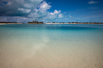 Golden color of the sand of shallow water on the beach of a tropical island with palms and clouds in a distance