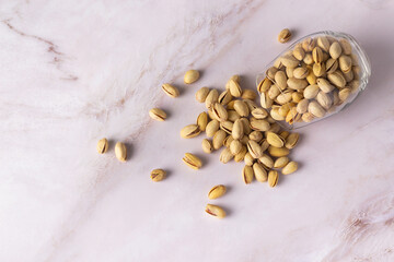 A glass jar has toppled, scattering roasted pistachios across a marble countertop, against a simple...