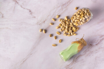 From above, you'll find a green bottle of pistachio milk positioned adjacent to a clear glass container brimming with shelled pistachios, all set against a backdrop of elegant white marble.