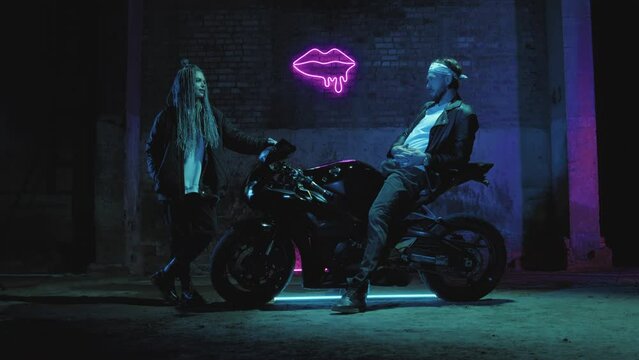 A guy on a super sport motorcycle stands talking to a girl against a pink neon sign