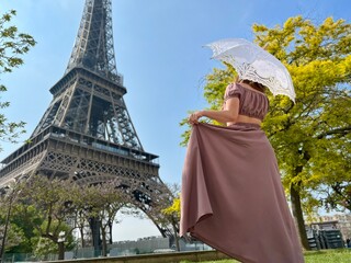 from the back we see a girl in a beautiful long brown dress in a retro style with an umbrella who walks towards the eiffel tower holding the dress with one hand. High quality photo