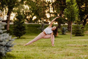 Young athletic woman practice yoga in green park on grass in summer morning. Stand in extended side angle pose in sportswear, forest background. Concept of healthy lifestyle and harmony with body.