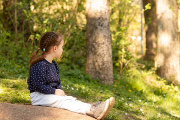 Little beautiful girl sits on a stone surface in a park on a blurred green natural background