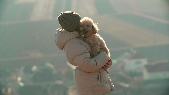 A young woman is kissing and hugging a beige toy puddle dog. She is holding the dog in her hands on a nature background. She is wearing jacket and cap. High quality FullHD footage