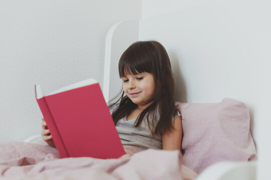 one todder girl 5 years old brunette with long hair sits in bed and reads a book and smile
