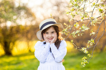 Portrait of adorable little girl in a white dress and hat, standing under flowering trees, in spring