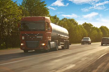 Tanker truck driving fast with other cars on a road with trees against a sky with clouds