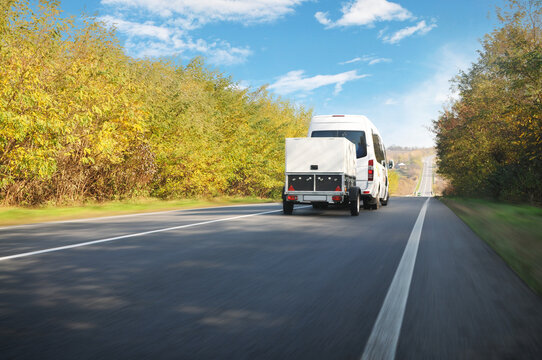 White van with a trailer driving fast on a countryside road against a sky with clouds