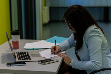 Businesswoman writing with pencil on notepad in the office with laptop and calculator on table