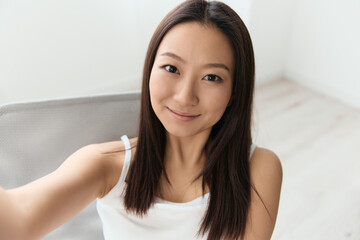 Smiling happy tanned lovely young Asian woman doing selfie sit on chair at home interior living room. Distance communication, Remote work, Social media concept. Cool offer Wide angle Selfie view