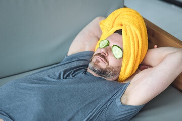 Handsome man lying on the sofa with cucumber slices on the eyes and yellow towel on the head. Concept of male self-care, skin care, relaxation at home