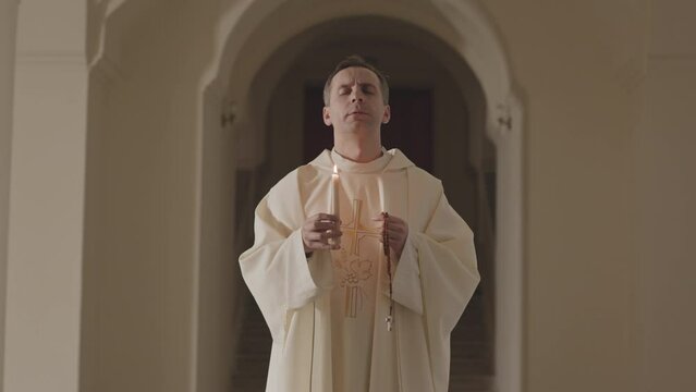 Medium slowmo of mature Caucasian priest in long white alb holding burning candle and rosary beads and praying to God, standing at Catholic church corridor