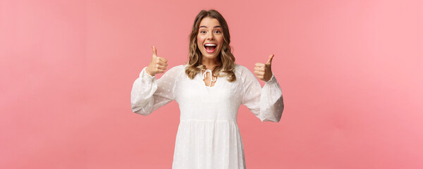Portrait of excited feminine blond girl in white dress, show thumbs-up in approval, like being...