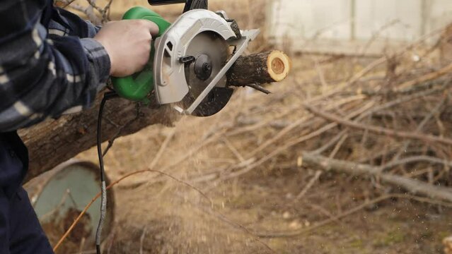 Super slow motion close-up of the hands of garden worker with manual electric circular saw sawing off chunk from the trunk of an apple tree.