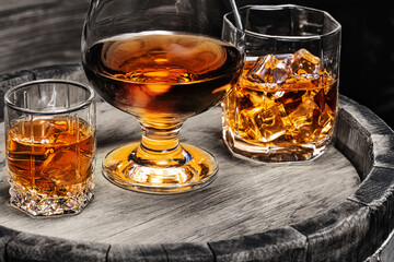 Whiskey, Cognac, Brandy, in glass of ice stand on wooden barrel of whiskey. Three glasses with alcoholic beverages whiskey, cognac, brandy close-up on wooden barrel.