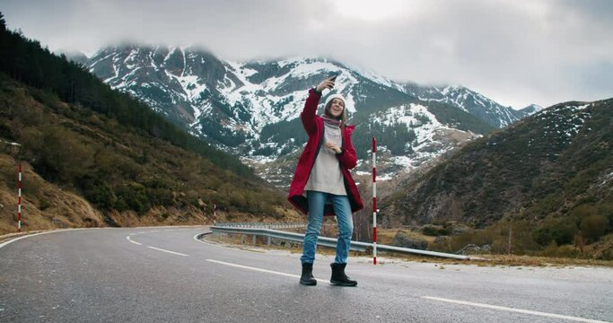 Young adult woman does a spin and laugh in camera. Traveler on empty mountain road makes selfie photo or video on smartphone for blog or social media. Cheerful and happy girl on vacations