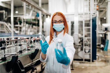 Beautiful female worker in workwear and with protective mask on her face working in bottling factory. Inspection quality control.