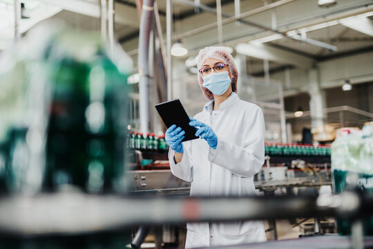 Beautiful Female Worker In Workwear, With Disposable Protective Cap And With Protective Mask On Her Face Working In Bottling Factory. Inspection Quality Control.