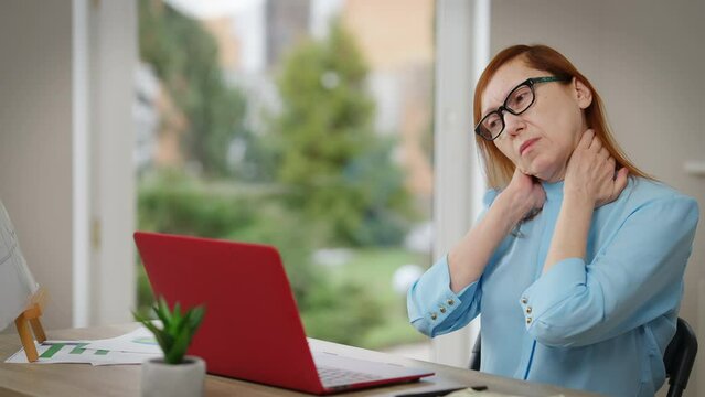 Sad tired mature woman stretching painful neck sitting at table with laptop. Portrait of upset Caucasian businesswoman massaging muscles indoors in home office. Sedentary lifestyle and health