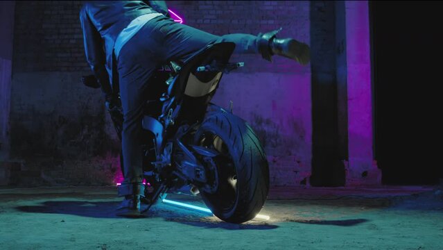 A male biker in a leather jacket sits on a sports bike against a neon pink signboard on a brick wall