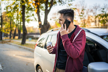 Young caucasian man standing by his car in autumn day or evening holding a cup of takeaway coffee looking to the side while waiting on the street in town copy space making a phone call talking