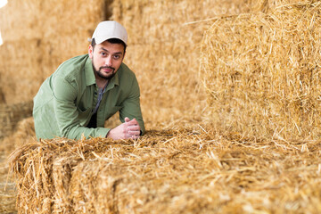 Naklejka na ściany i meble Portrait of positive young adult man farmer posing at hay storage on dairy farm