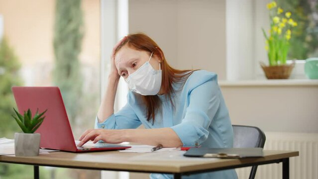 Portrait of exhausted tired mature woman in Covid-19 face mask messaging online on laptop caughing. Ill Caucasian freelancer working online in home office on pandemic quarantine