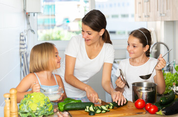 Cheerful woman with two little daughters prepare vegetable salad in the kitchen at home