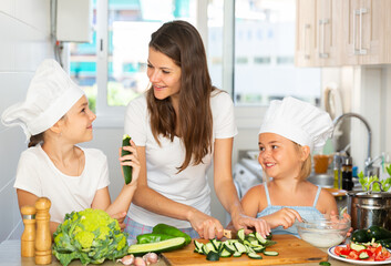 Woman with two kids in ches hats cooking together cutting and tasting vegetables for salad at home kitchen
