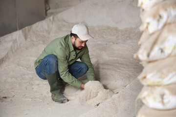 Confident focused young bearded farmer holding handful of corn meal in warehouse of livestock farm, examining organic ingredient for use in animal feed