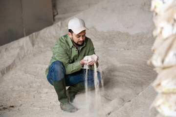 Confident focused young bearded farmer holding handful of corn meal in warehouse of livestock farm, examining organic ingredient for use in animal feed