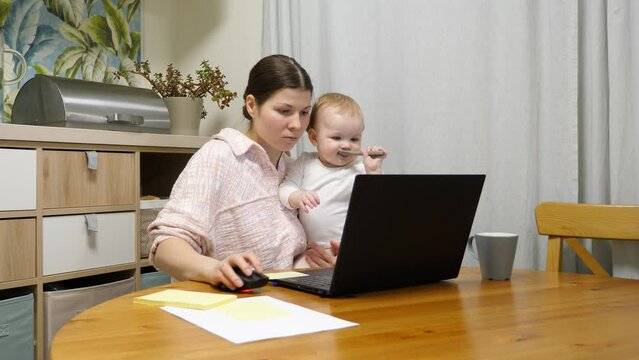Young woman works part-time on internet while staying home with young daughter during her maternity leave. She uses laptop, holds her small child in arms, and at same time does her work assignments