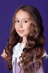 A cheerful little child, smiling and feeling happy. Cute little girl in the studio in a lilac dress and with long hair on a purple background.