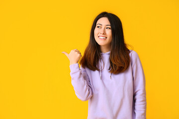 Inviting young Asian woman pointing at something on yellow background