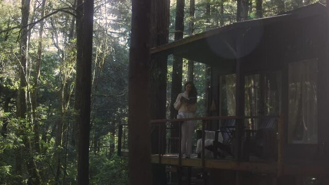 Young woman reading a book and drinking hot tea on a porch of a cozy cabin in the woods. Log cabin in the middle of woods sunny wooden bright mountain cabin rustic house architecture. 