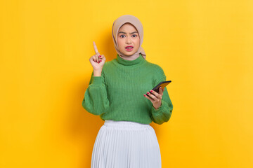 Shocked beautiful Asian woman in green sweater holding mobile phone and pointing fingers up having a good idea isolated over yellow background