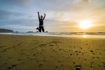Adventurous athletic woman jumping in the air and smiling at the camera on a Pacific Northwest beach during a beautiful sunset.