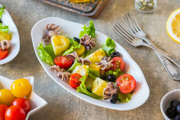 Potato salad with octopus, tomato and leaf lettuce on a gray oval plate on a gray concrete background. Mediterranean, Italian cuisine.